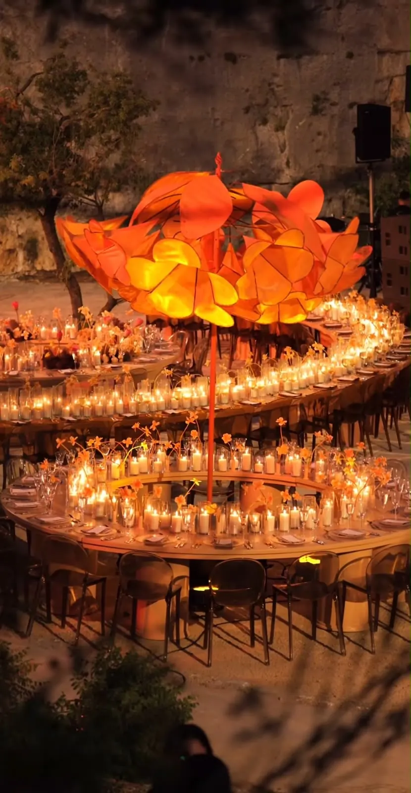 The glowing orange blossom tree above a ring of candlelit wedding tables at night